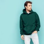 Handsome confident hipster  model.Sexy unshaven man dressed in summer stylish green hoodie and jeans clothes. Fashion male with curly hairstyle posing in studio. Isolated on blue